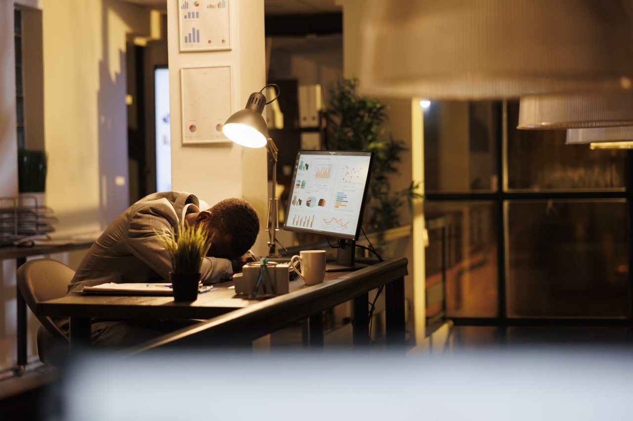 Funcionário cansado dormindo sozinho na mesa do escritório, enquanto a tela do computador está acesa com gráficos.