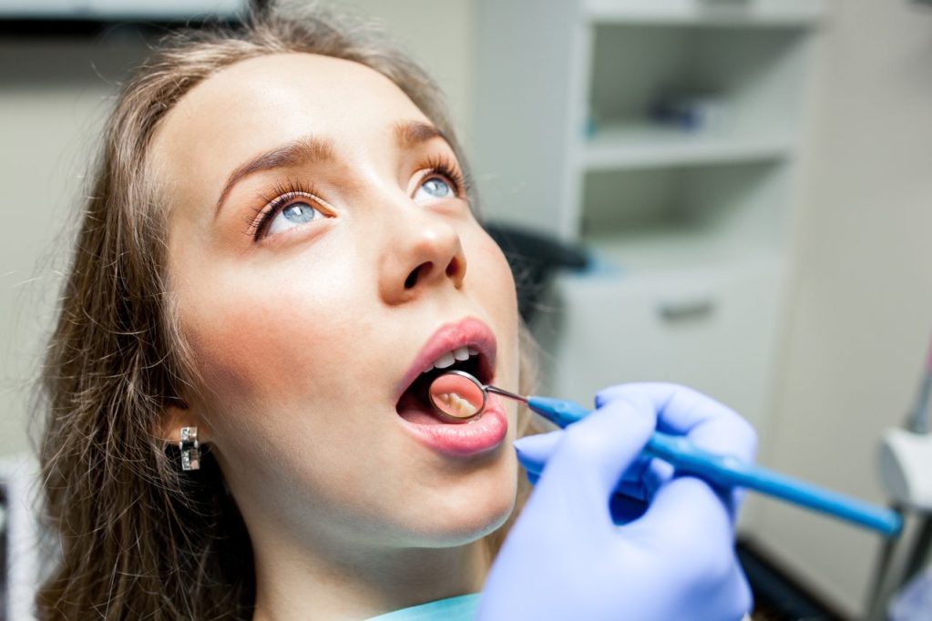Mulher jovem em consultório de dentista. O dentista olha seus dentes com espelhinho.
