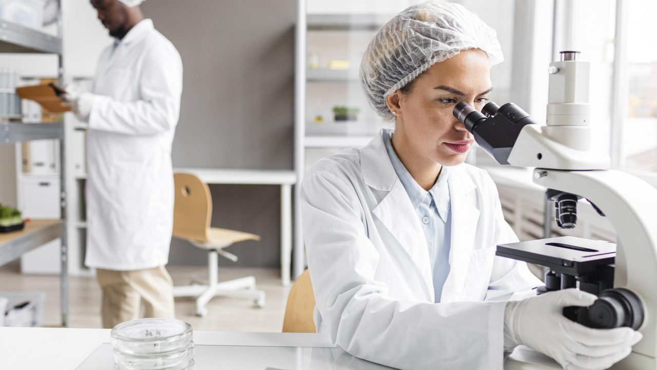 Mulher em laboratório olhando microscópio, ao fundo tem um homem escrevendo algo em prancheta