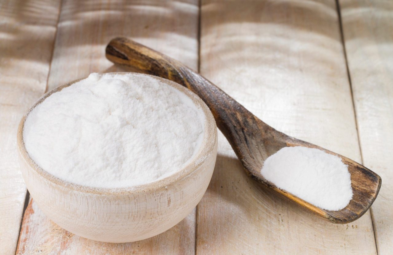 Cumbuca cheia de pó de bicarbonato e ao lado uma colher de pau com um pouco de bicarbonato.