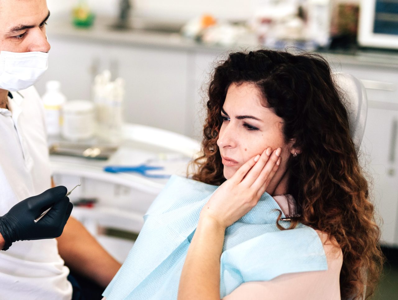 Paciente no dentista. Ela está sentada na cadeira odontológica com a mão na bochecha e expressão de dor. Enquanto isso o dentista está ao seu lado prestando atenção e segurando um instrumento odontológico.