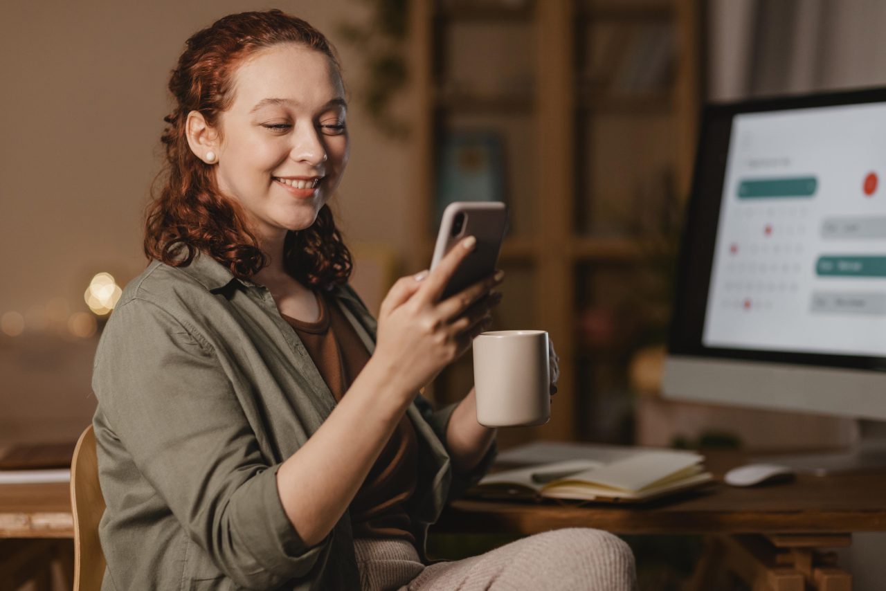Mulher ruiva sentada em escritório segurando celular e xícara de café. Ela está sorrindo e na sua frente tem um computador com um calendário aberto na tela.