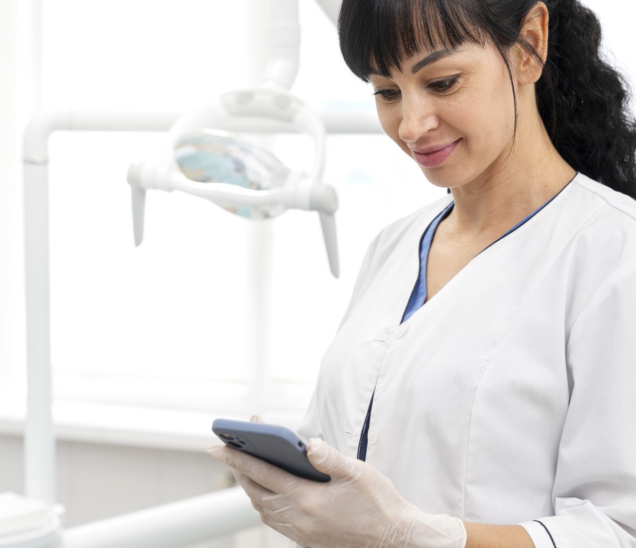 Doutora usando jaleco branco em consultório odontológico, enquanto olha sorrindo seu celular.