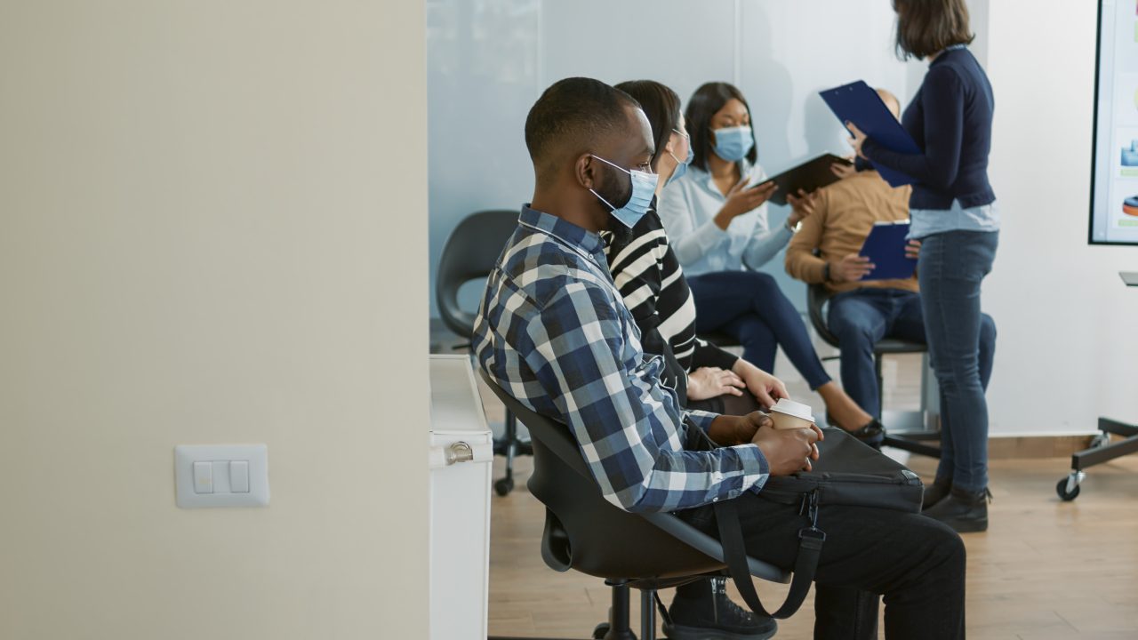 Pacientes sentados na recepção de clínica. Todos estão usando máscaras descartáveis azuis. Em pé tem uma recepcionista entregando pranchetas para eles preencherem seus dados.