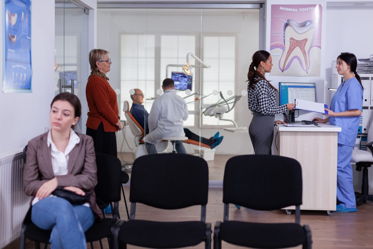Espaço de clínica odontológica amplo. É possível ver uma paciente sentada na recepção, enquanto outras duas pacientes estão em pé para preencher ficha com a recepcionista. Ao fundo, tem o consultório odontológico com portas de vidro, sendo possível ver o dentista e o seu paciente.