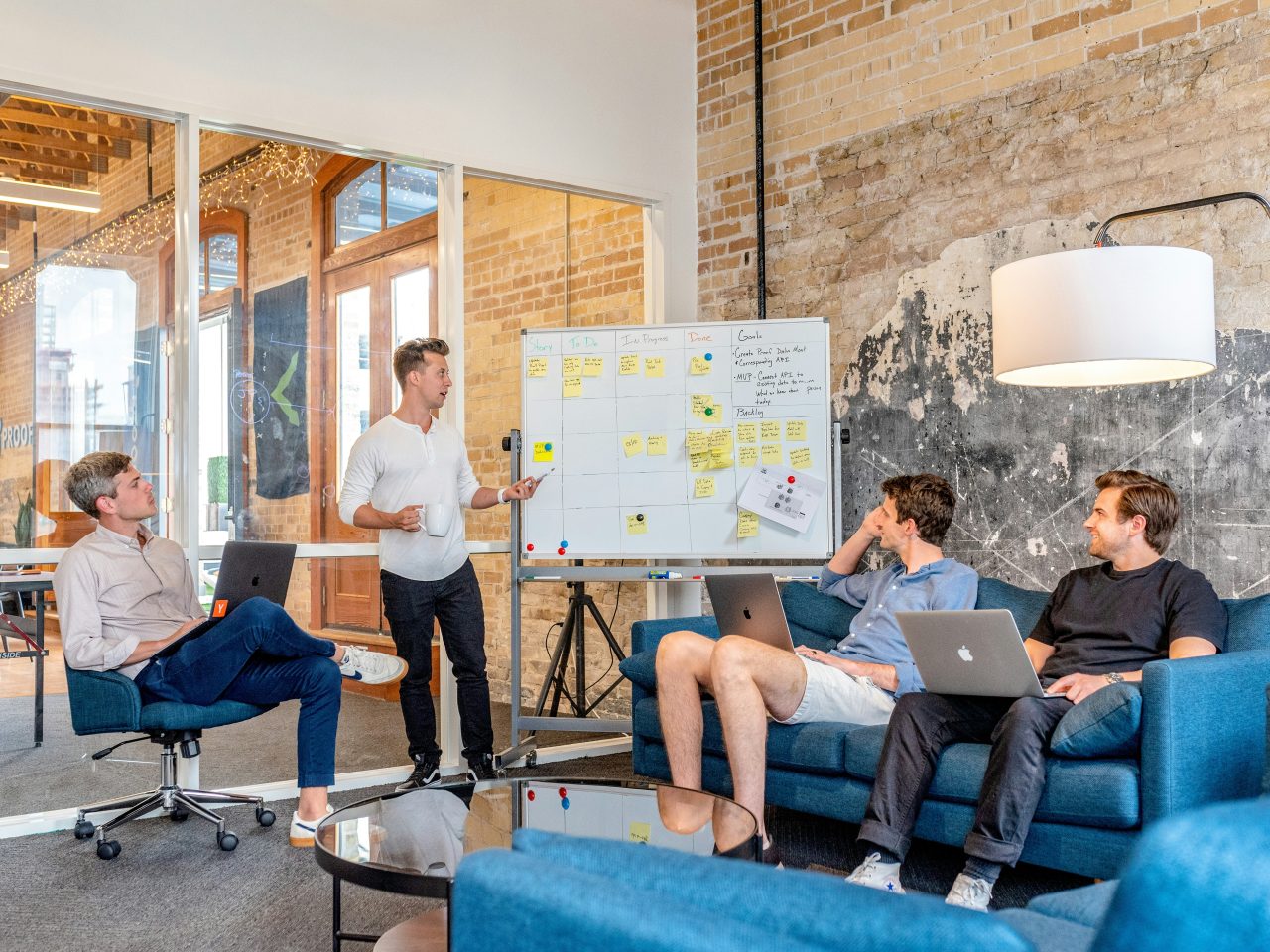 Sala de reuniões de empresa. Há homens de roupas casuais sentados nos sofás azuis. Em pé tem um homem explicando planejamento que está em quadro com post-its.