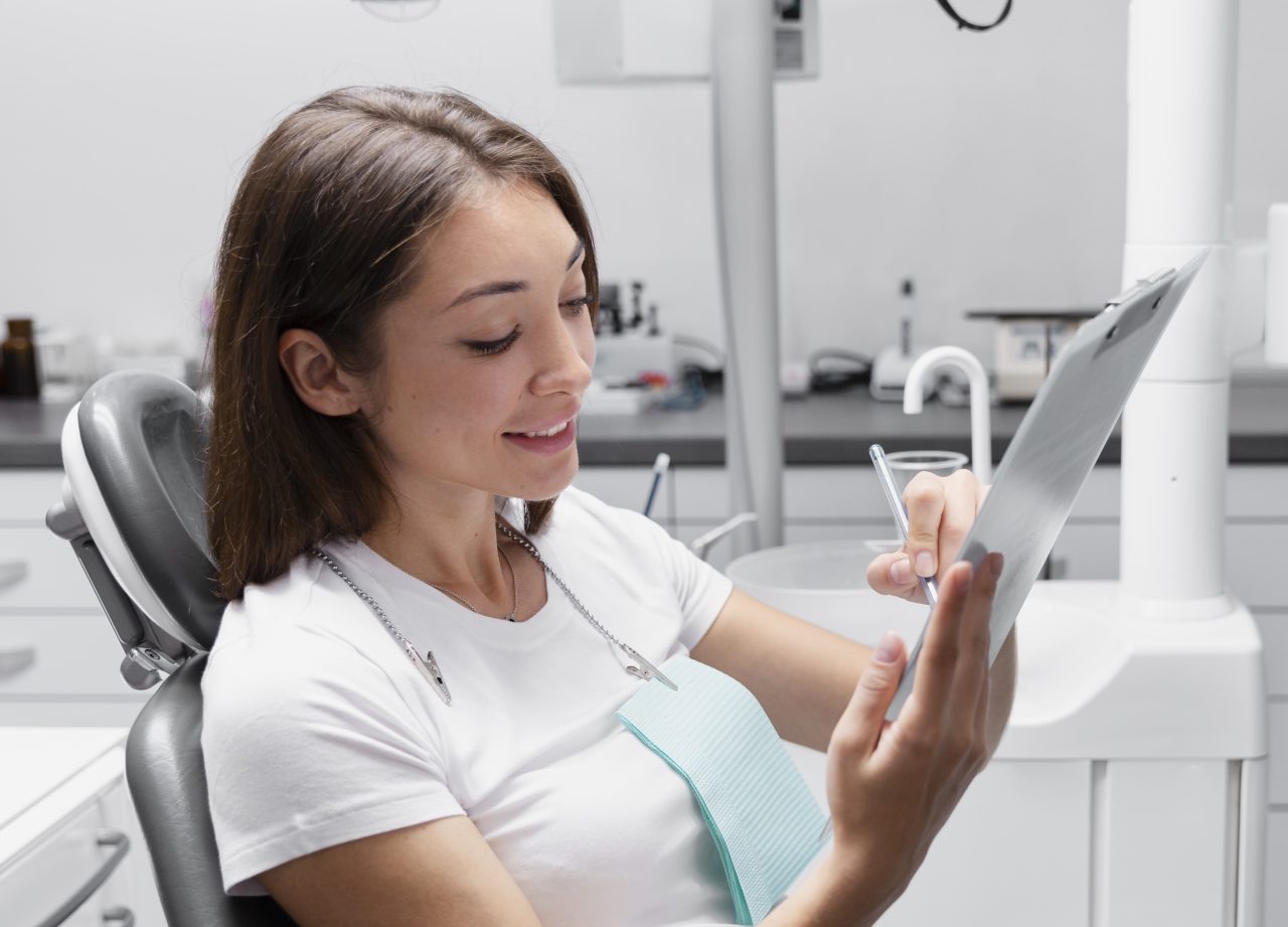 Paciente mulher de cabelos médios castanho está sentada em cadeira odontológica, dentro de consultório inteiramente branco. Ela segura uma prancheta nas mãos enquanto preenche seus dados com caneta.