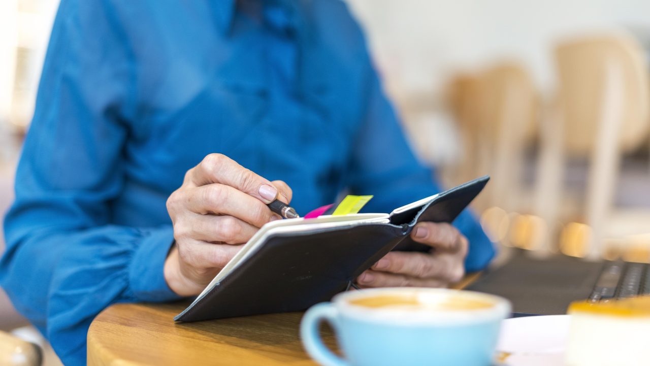 Pessoa de camisa azul está anotando algo em agenda, em sua frente tem uma xícara de café.