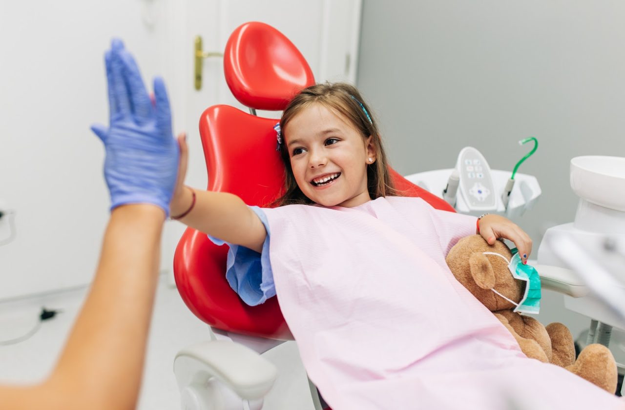 Menina segurando ursinho com uma mão, e com a outra batendo high five com dentista. A criança está num consultório de dentista.