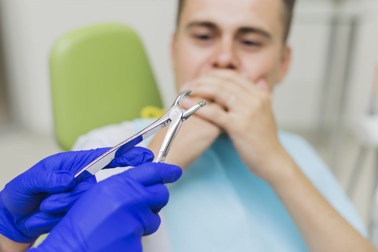 dentista segurando instrumento para extração de dente, enquanto isso o paciente está olhando com medo e cobrindo a boca com a mão
