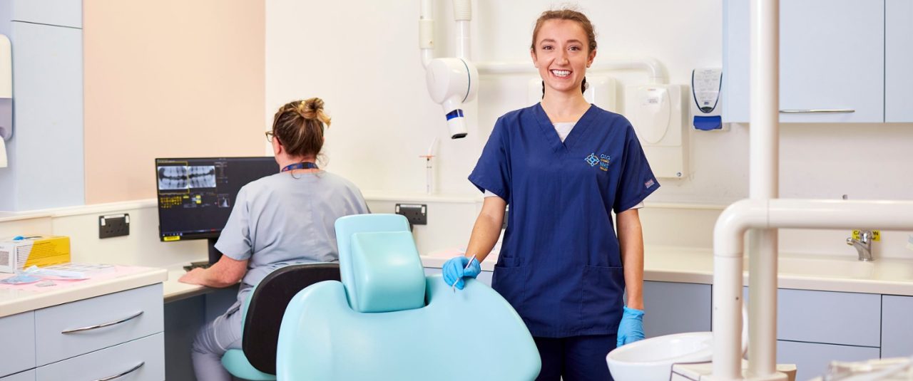 Dentista em pé sorrindo ao lado da cadeira odontológica vazia e assistente está sentada no computador olhando raio-x.