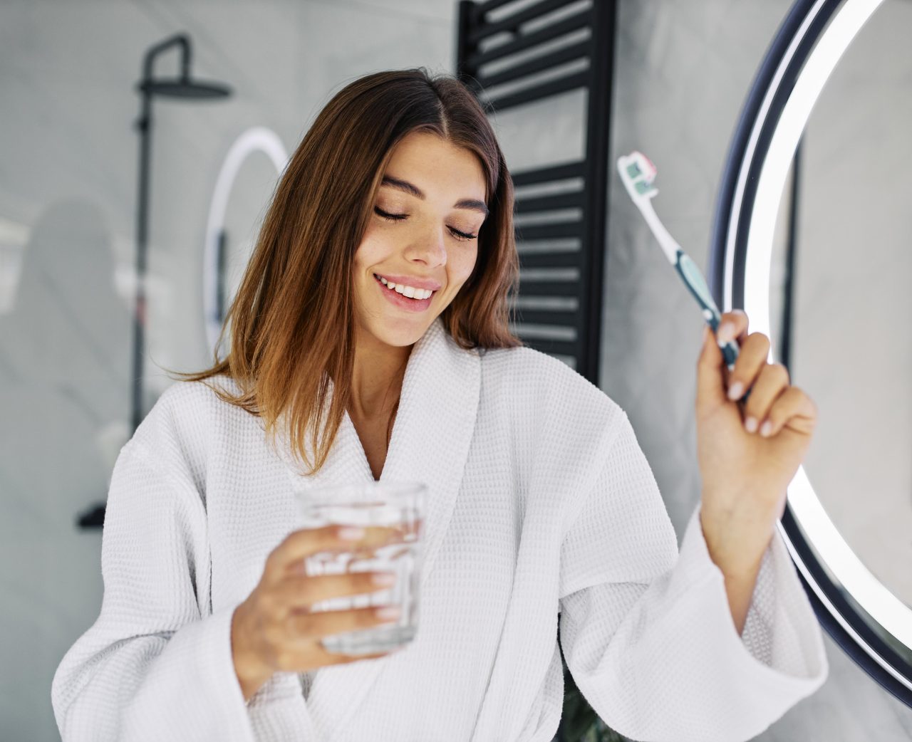 Mulher feliz no banheiro de roupão branco, segurando sua escova de dentes e um copo de água.
