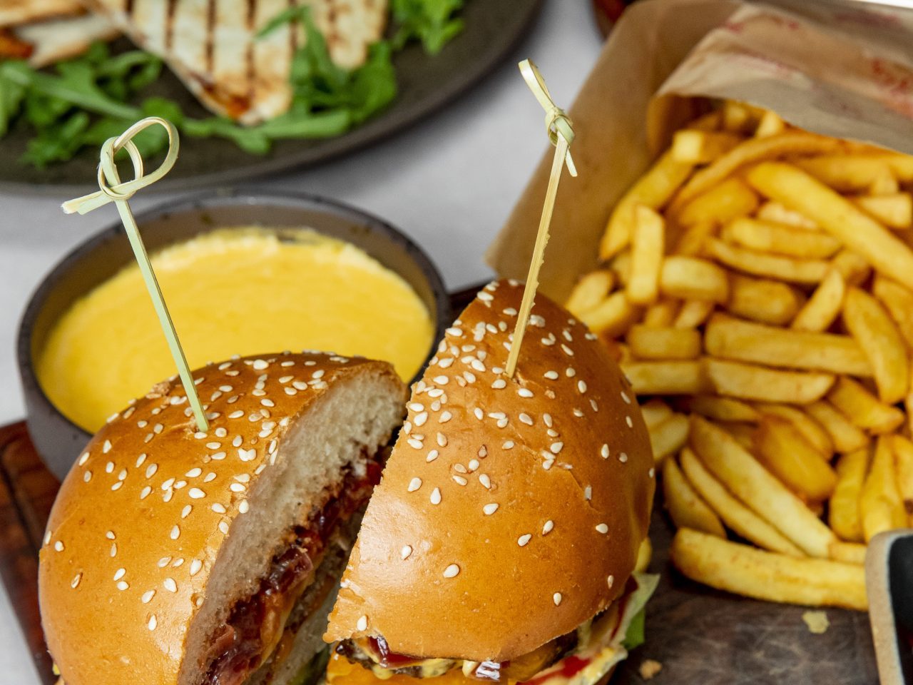 Foto de diversos alimentos. Na frente tem um sanduíche de fast food, um fondue de queijo atrás, batata frita, e no fundo um sanduíche natural.