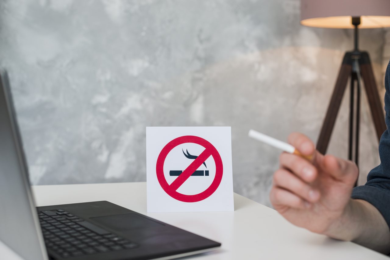 Homem sentado em frente a notebook segurando um cigarro, ao seu lado na mesa tem uma plaquinha de proibido fumar