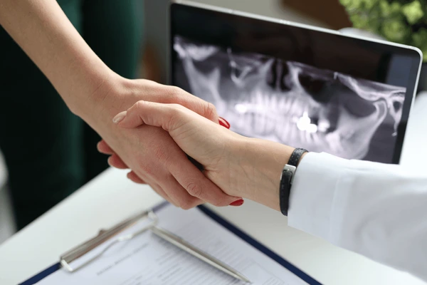 Dentista e paciente dando as mãos. No fundo é possível ver raio-x da arcada dentária em um tablet.