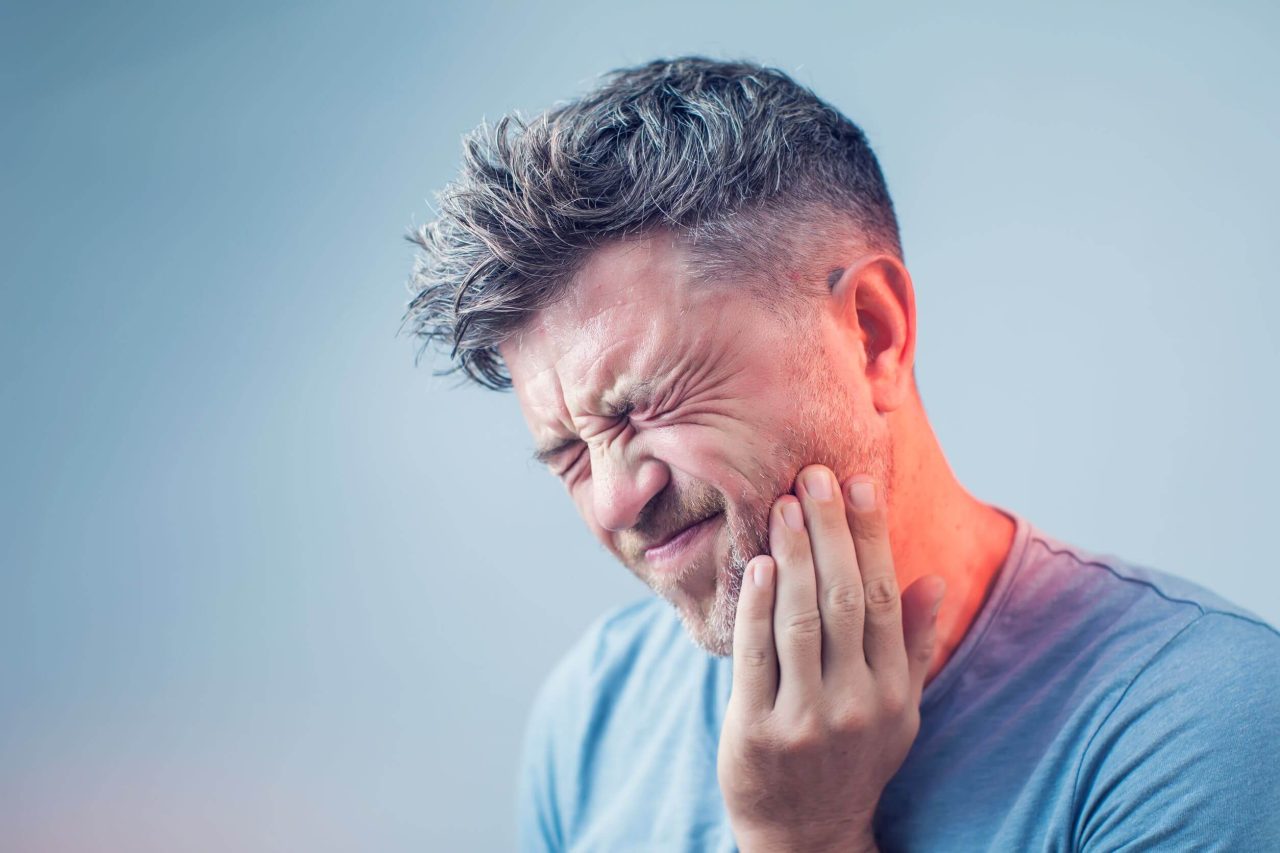 Homem de cabelos grisalhos e usando camisa azul está com dor de dente, seu rosto expressa a dor por estar todo contraído e ele apoia os dedos na bochecha.