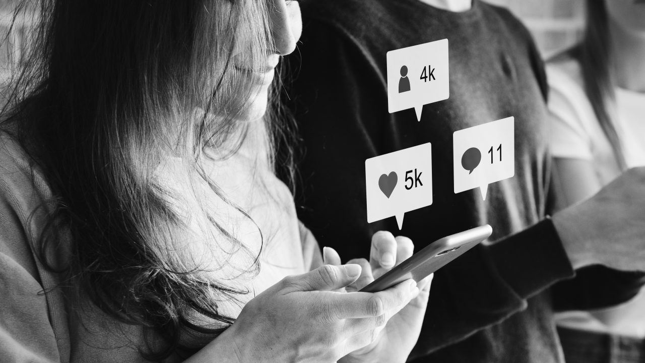 Mulher jovem usando o celular, da sua tela está mostrando o número de curtidas (5k), comentários (11) e seguidores (4k). Ao seu lado tem outros jovens em pé encostados na parede de tijolos.