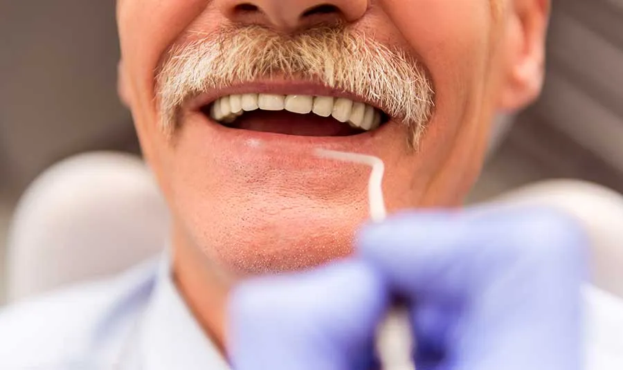 Senhor idoso e de bigode grisalho está sorrindo. Ele possui implantes e está no dentista.