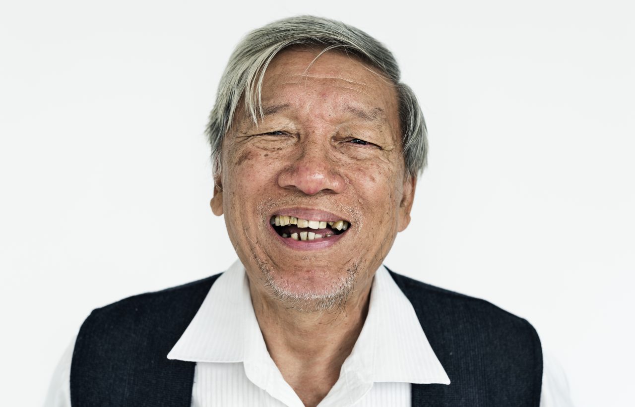 Idoso de origem asiática, com cabelos grisalhos e usado suéter preto está sorrindo e seus dentes são tortos e desgastados.