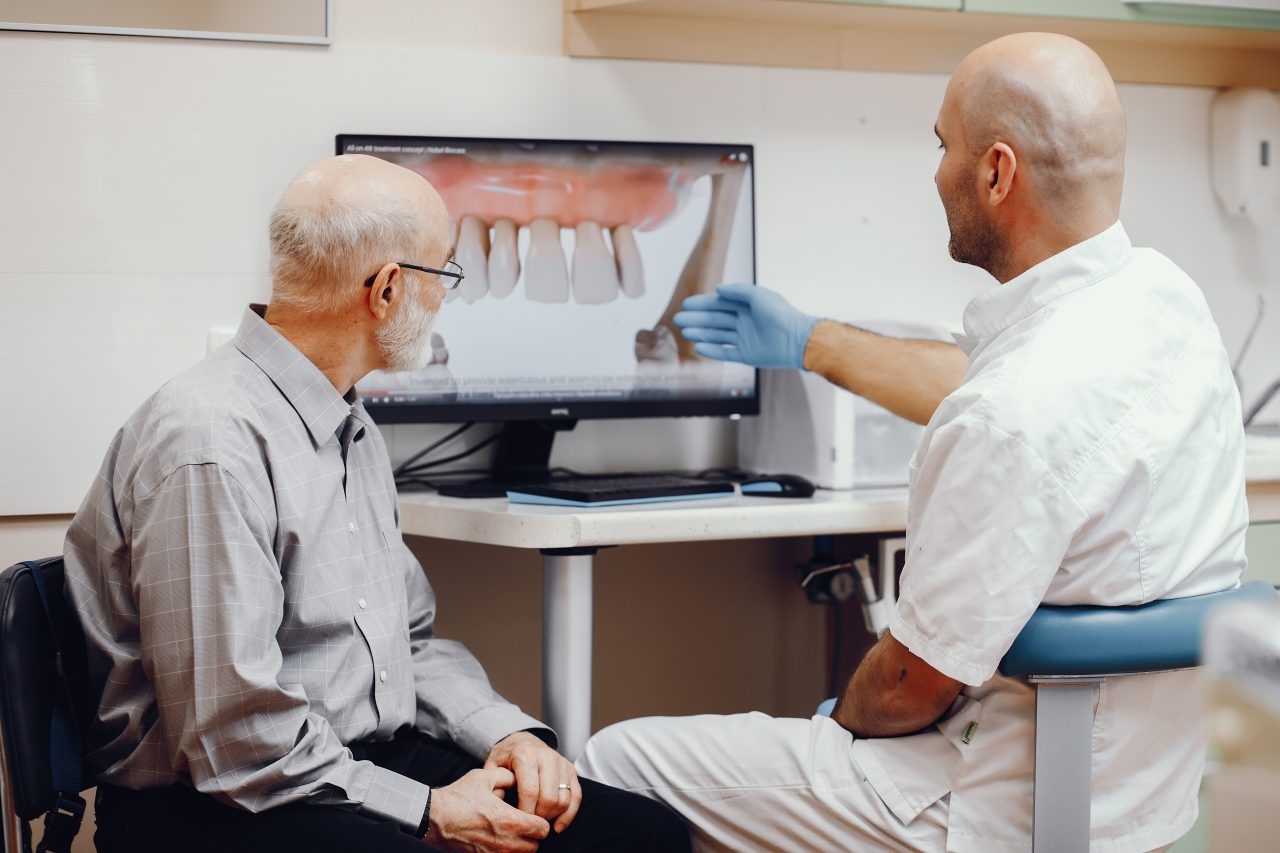 Dentista explicando planejamento do tratamento para senhor idoso, mostrando seus dentes na tela do computador.