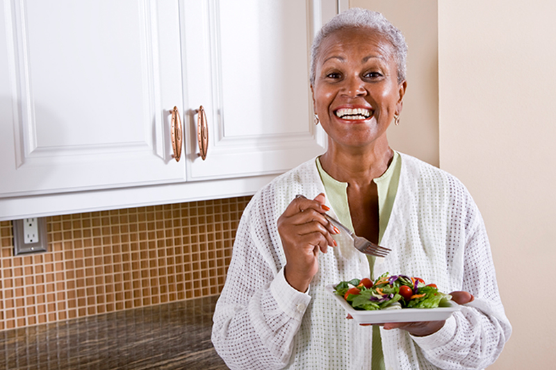 Senhora idosa negra de cabelos grisalhos curtos está comendo salada em pé na cozinha, ela sorri para foto.