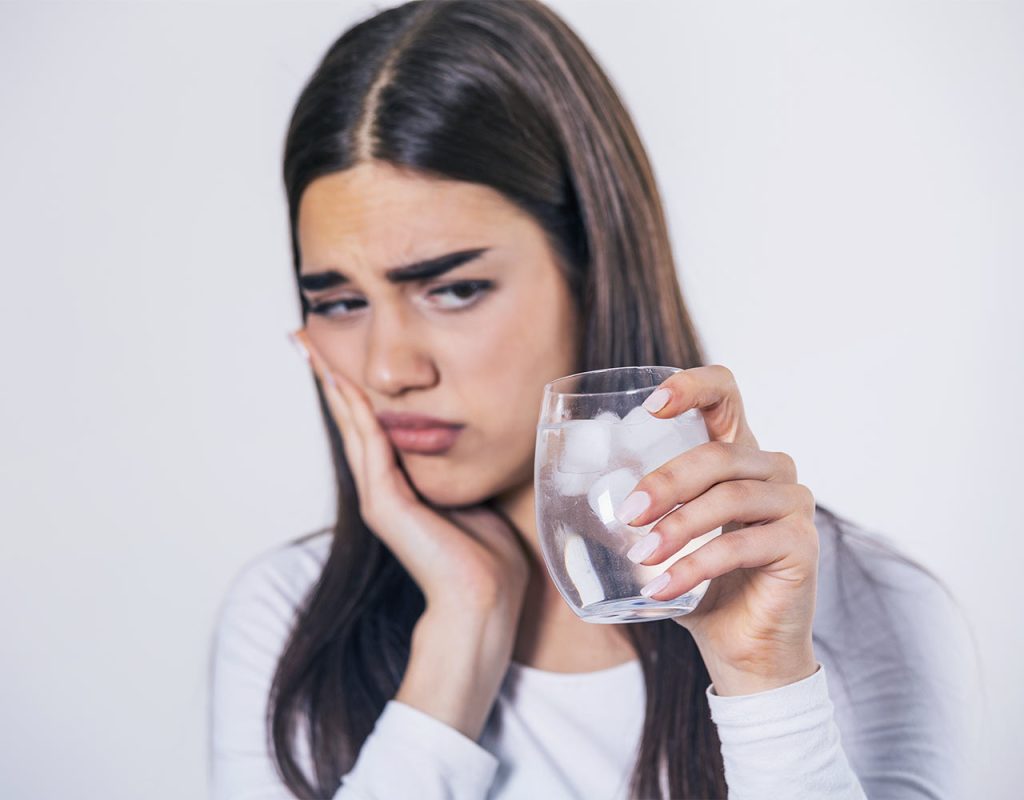 Jovem mulher encarando com expressão de dor copo de água com gelo enquanto aperta a bochecha