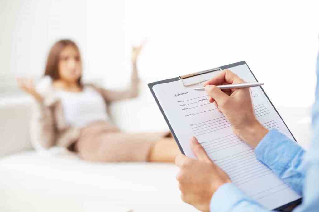 Psicóloga está com uma prancheta para anotações e ao fundo desfocado uma mulher está no sofá desabafando.