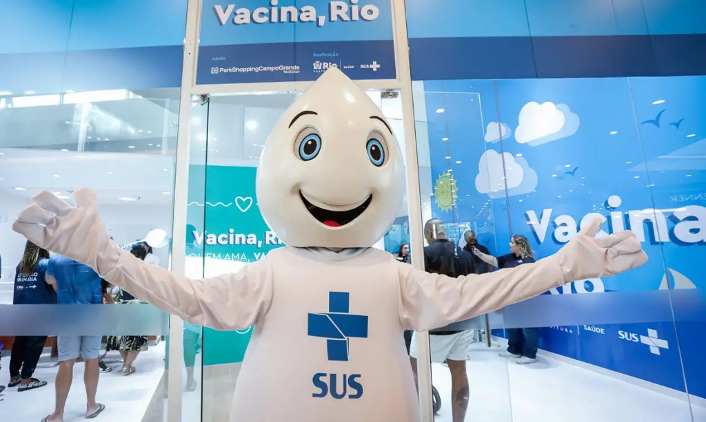 Pessoa vestida de Zé Gotinha com logo do SUS. Está em frente a uma unidade do SUS de vacina no shopping.