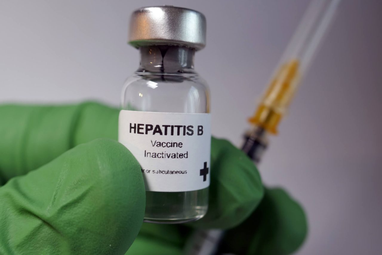 Pessoa de luva verde segurando frasco de Vacina contra hepatite B e seringa.