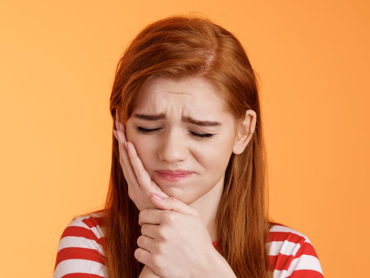 Garota ruiva com camiseta listrada está a palma da mão apoiada na bocheça. Ela está com expressão de desconforto indicando dor de dente.