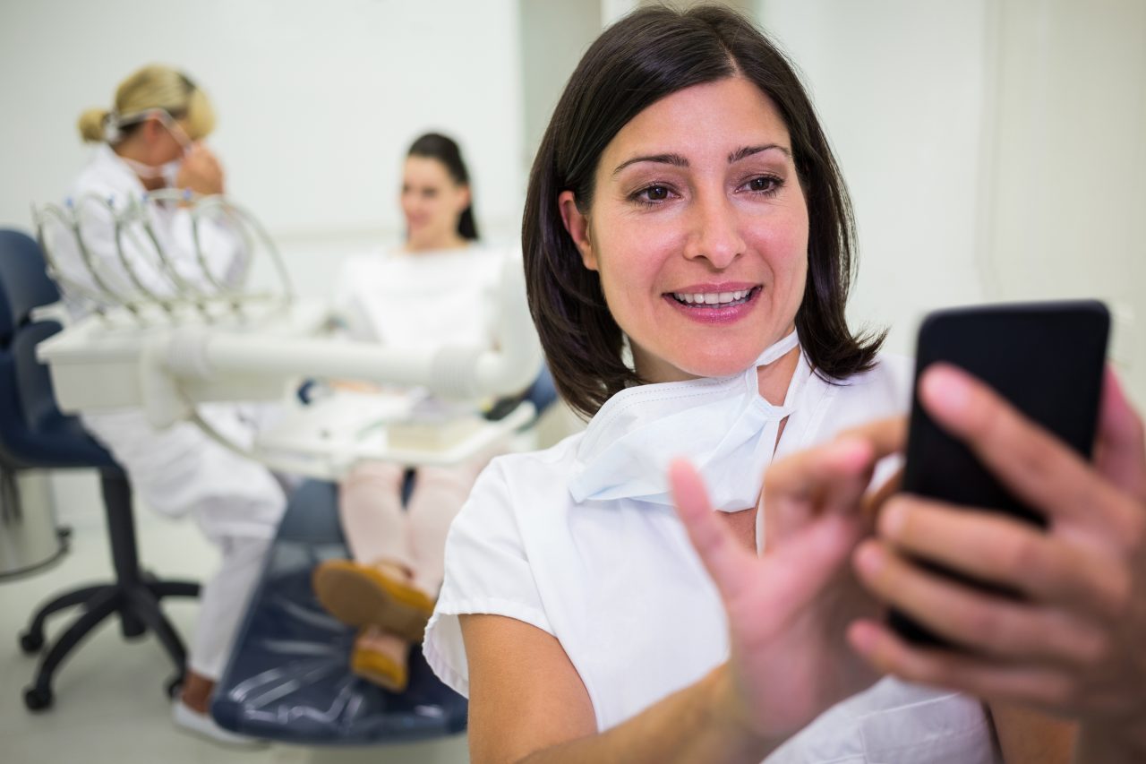 Assistente de dentista está checando mensagens em seu celular. No fundo dentista atende paciente.