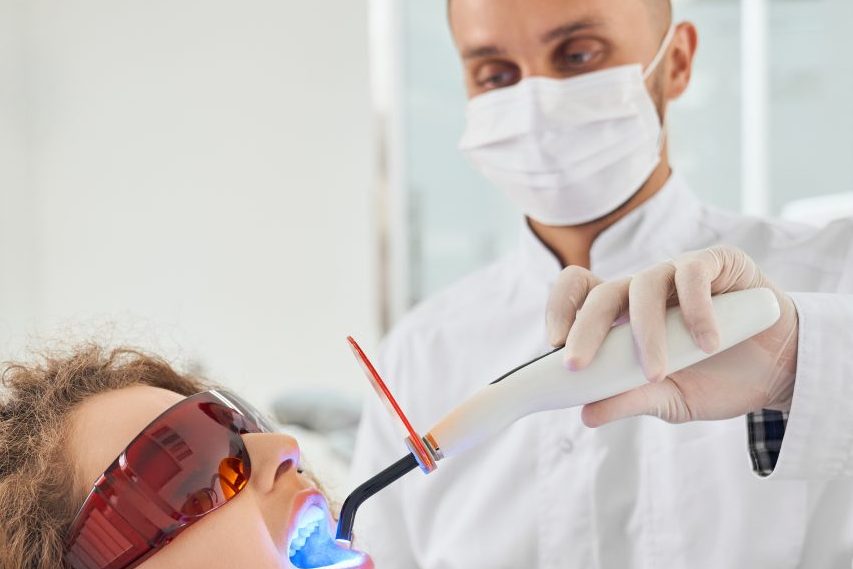 Paciente no consultório odontológico usando óculos de proteção enquanto dentista segura luz ultravioleta em seus dentes.