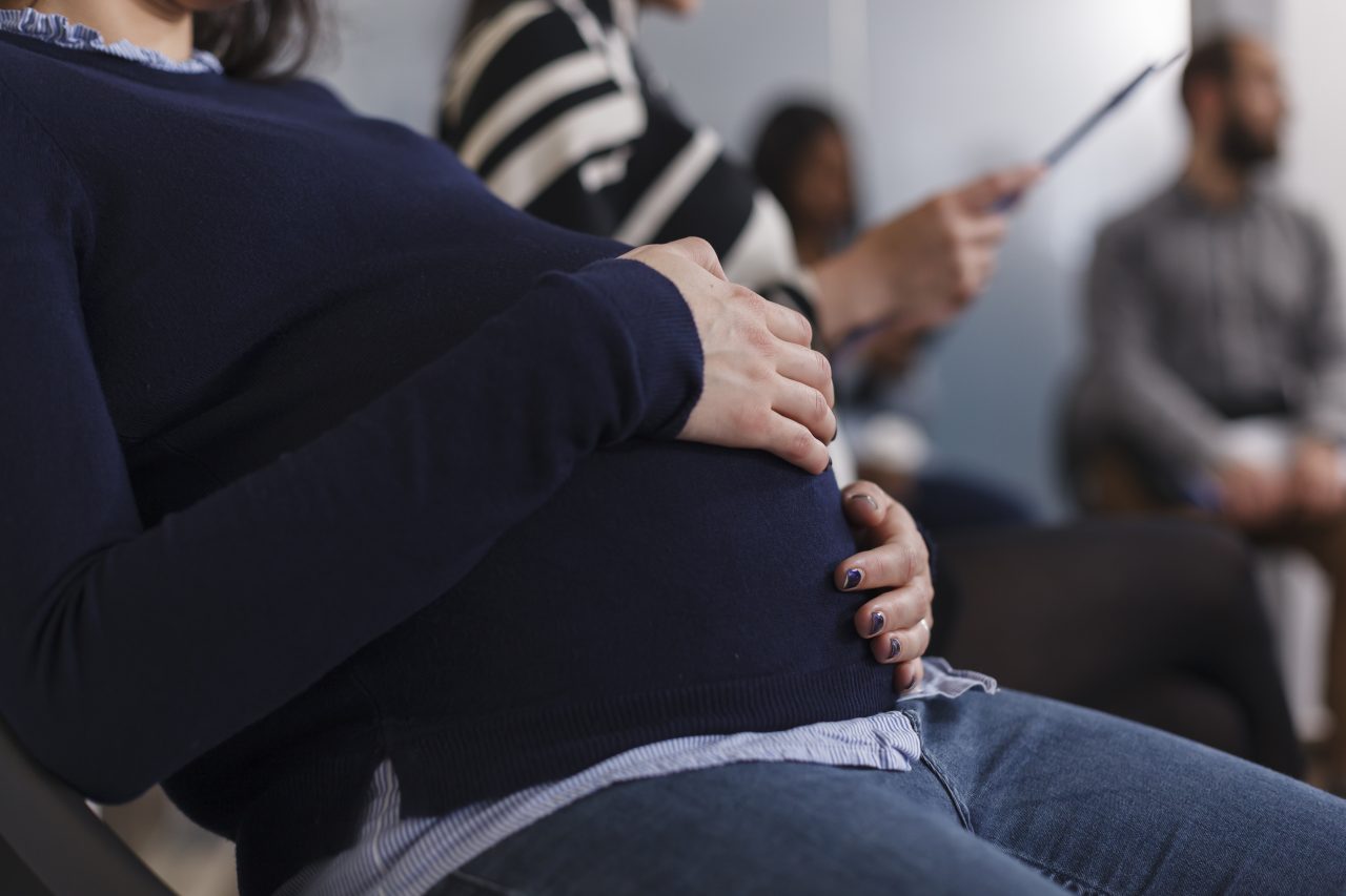 Na imagem, é uma clínica com pessoas sentadas na recepção. A pessoa mais perto da câmera é uma mulher grávida com as mãos na barriga.