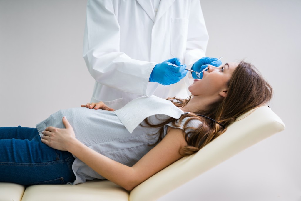 Dentista tratando dentes de jovem mulher grávida, deitada na cadeira odontológica.