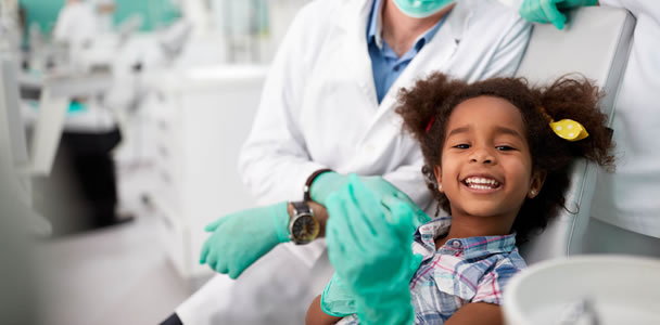 Odontopediatra usando jaleco ao lado de criança no dentista que está sorrindo e colocando luvas.