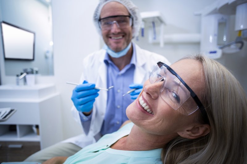Dentista está segurando materiais odontológicos, e sorrindo, enquanto paciente idosa olha para a câmera e também sorri.