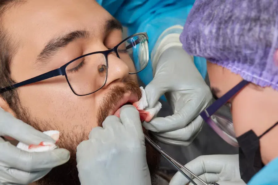 Dentista e assistente fazendo cirurgia bucal em paciente, e colocando gazes pois está sangrando.