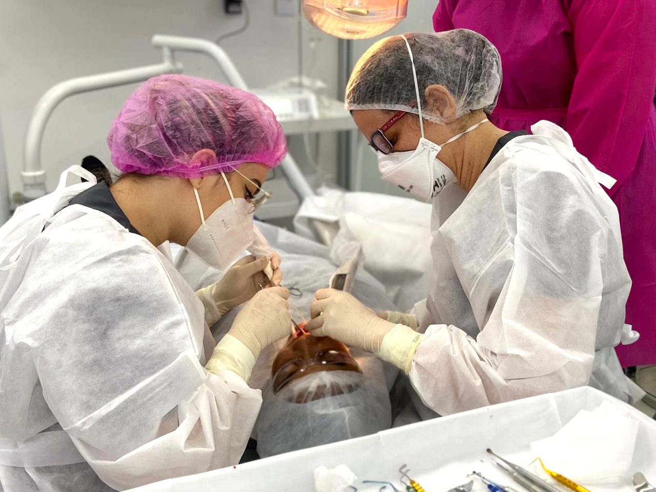 Duas alunas da EAP Goiás durante aula prática na clínica-escola com paciente. Ao fundo a professora está parada observando.