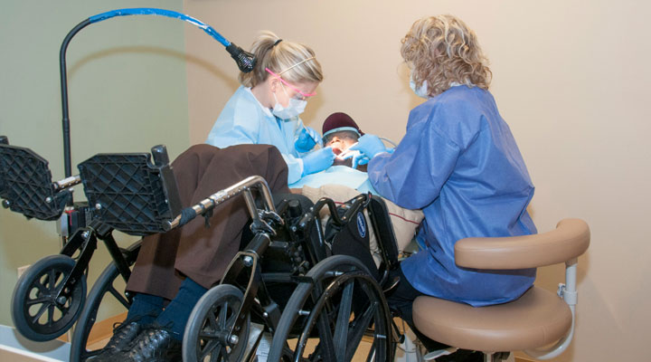 Dentista e assistente realizando tratamento odontológico em paciente cadeirante. A cadeira está deitada na mesa.