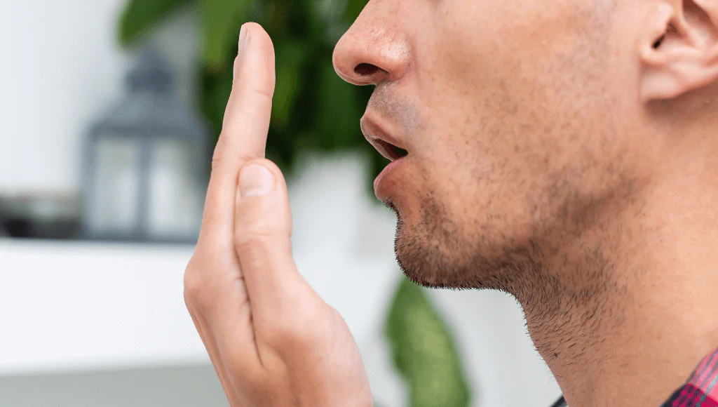 Homem soprando na própria mão para checar seu hálito. O fundo está desfocado.