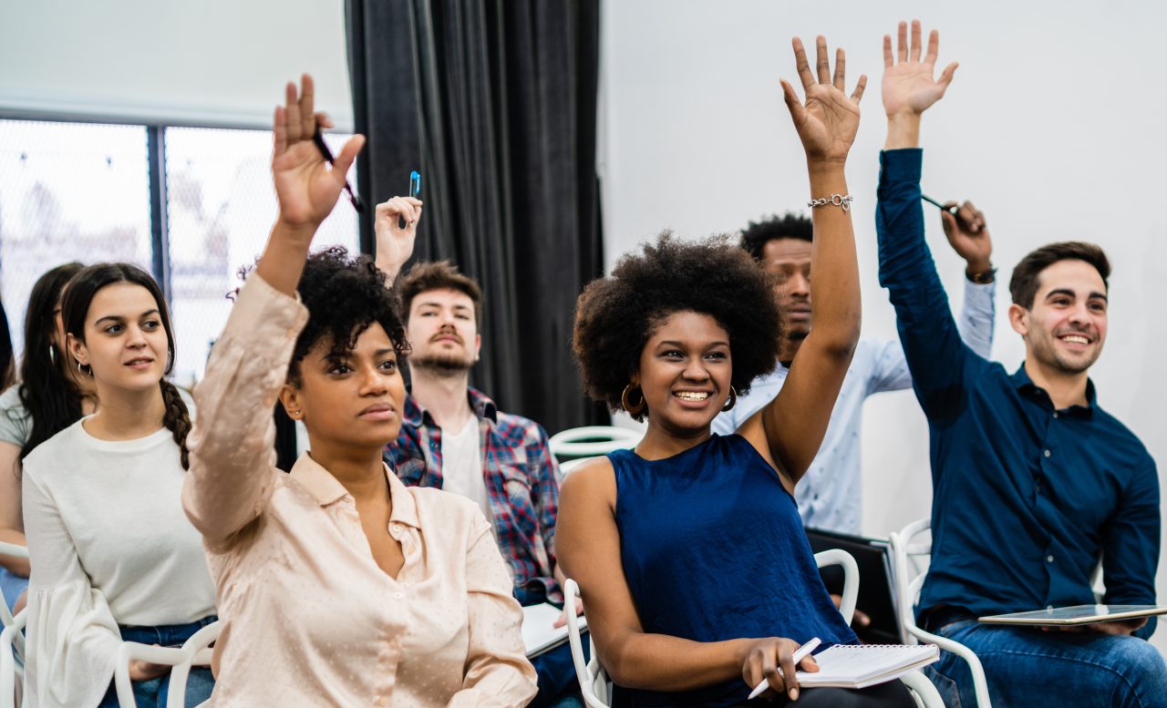 Pessoas em sala ouvindo palestras, algumas estão com as mãos levantadas para fazer perguntas e estão sorrindo.
