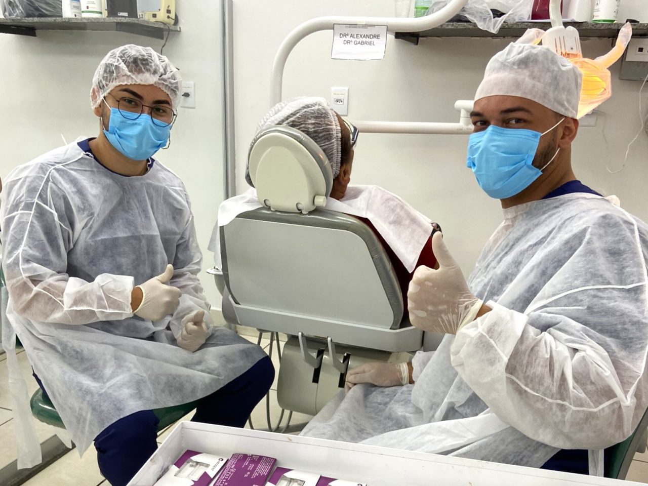 Dois alunos da EAP Goiás fazendo sinal de positivo com o dedo; Eles estão sentados ao lado da paciente.
