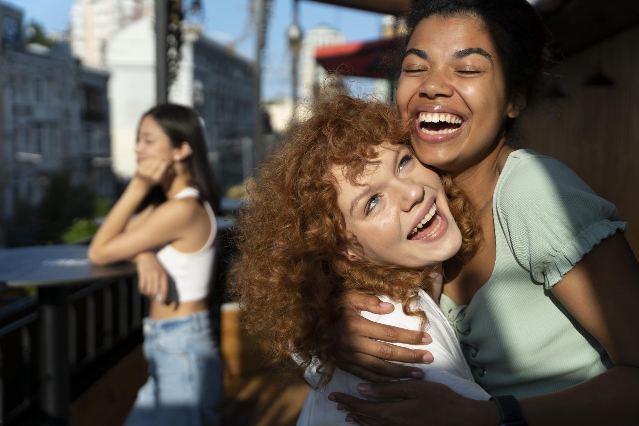 Duas garotas novas se abraçando e rindo muito. No fundo, tem desfocada uma garota com expressão triste olhando a cidade sozinha.