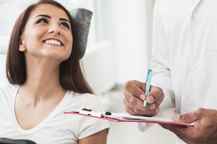 Dentista em pé ao lado de paciente anotando informações na prancheta, ela está sorrindo para ele.