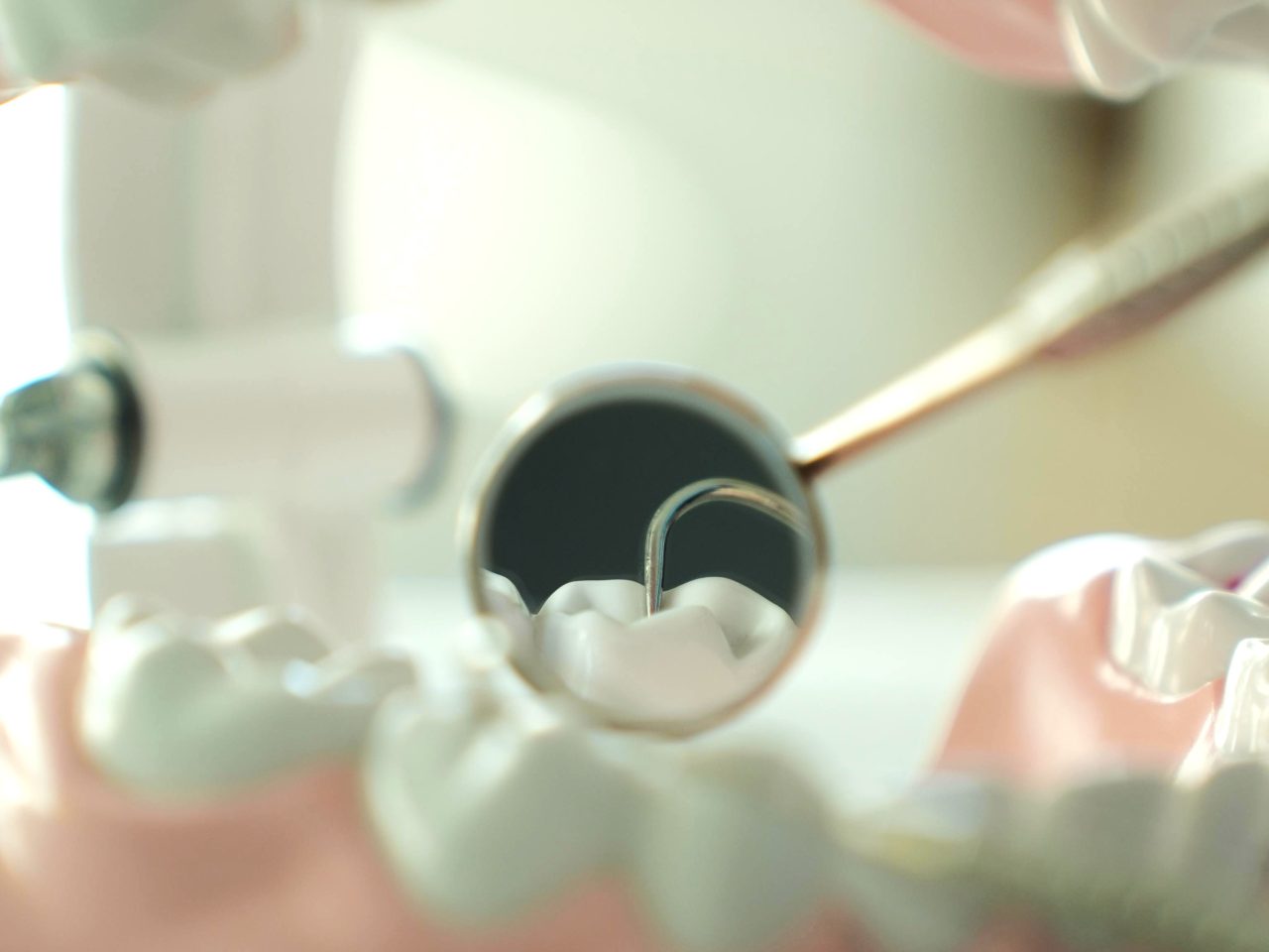 Imagem de um espelhinho de dentista olhando dente. A boca é um plástico, um manequim.