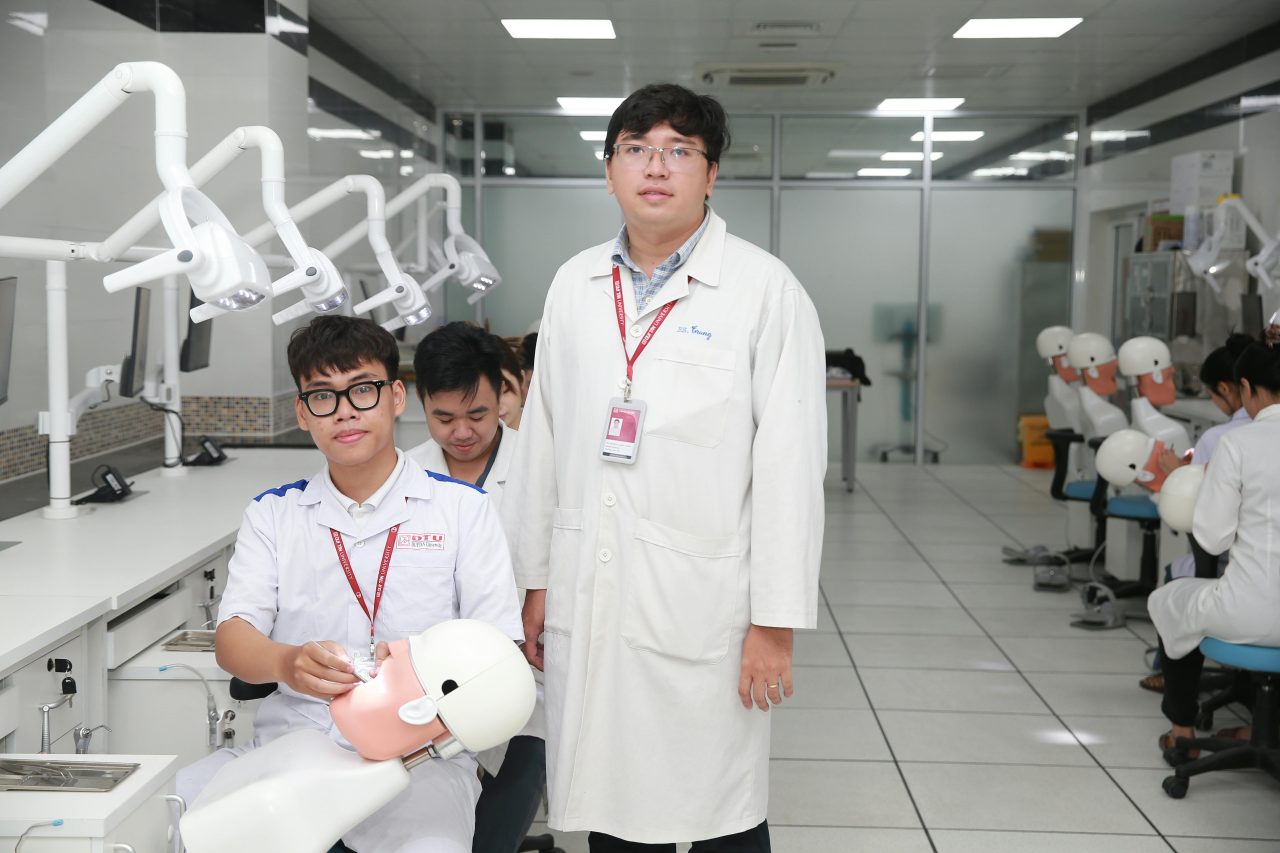 Professor ao lado de aluno na sala de odontologia em que estão aprendendo me bonecos.