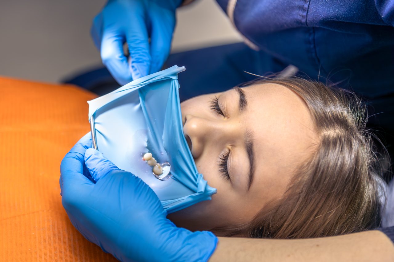 Jovem moça está no consultório odontológico sendo atendida por uma dentista que está colocando isolamento absoluto em sua boca.