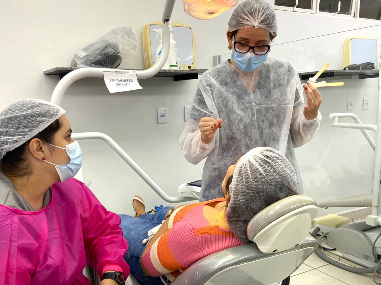 Dentista da EAP Goiás explicando procedimento para paciente. A doutora está em pé olhando para a paciente na cadeira, e tem uma assistente sentada do lado.