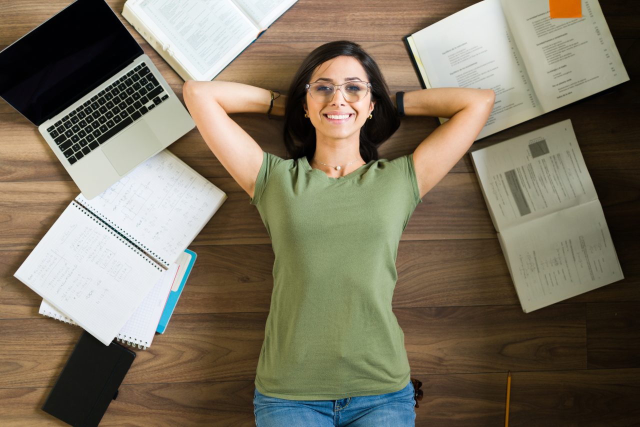 Mulher adulta está deitada no chão de madeira. Ao seu redor tem livros, cadernos e um notebook abertos. Ela está sorrindo e com as mãos atrás da cabeça;