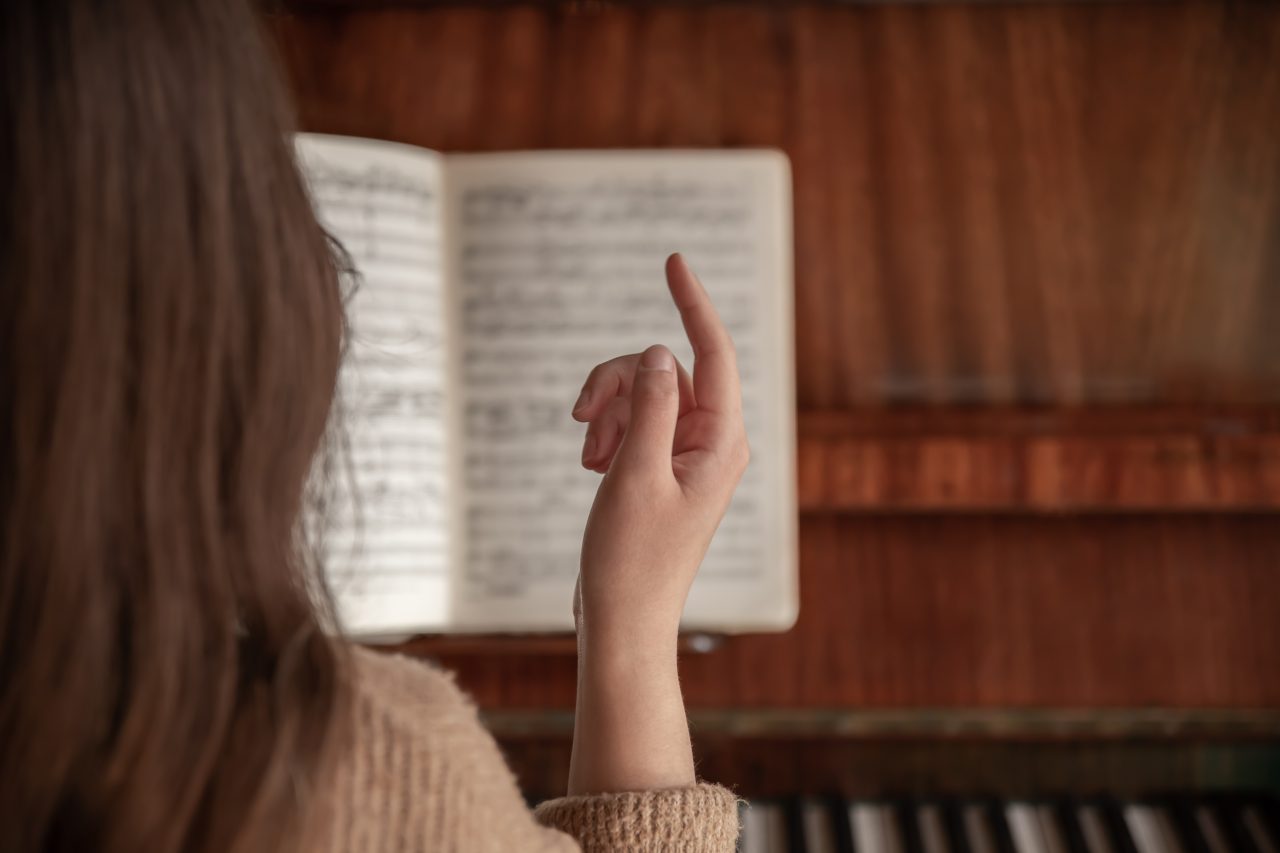 Jovem mulher sentada em frente a piano com a partitura aberta, ela está com o dedo levantado.